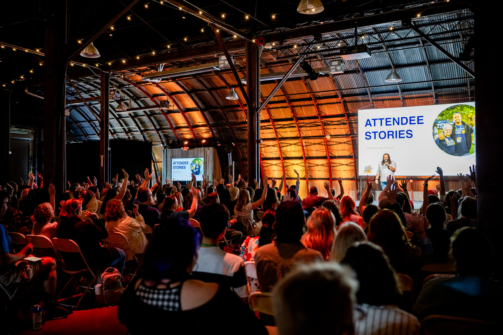 A packed room of people at NeuroDiversion 2026 in Austin, hands raised toward a speaker under warm string lights.